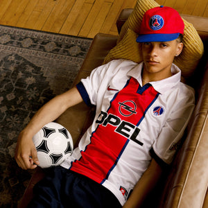 Man lying on a brown couch and holds a football. He wears the Paris Saint-Germain Swingman Snapback hat and a PSG jersey.
